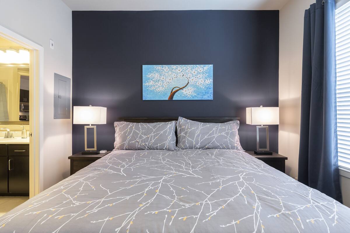 A bedroom with a bed, two bedside tables with lamps, a painting on the wall, and a window with curtains.
