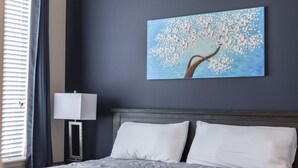 Ein Schlafzimmer mit einem Bett, weißen Kissen, einem Nachttisch mit Lampe und einem Baumgemälde an der Wand.