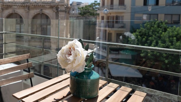 Ein Balkon mit einer hölzernen Bank und einer Vase mit einer weißen Blume. Im Hintergrund sind Gebäude und eine Kirche zu sehen.