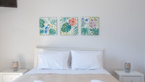 Ein ordentlich bezogenes Bett mit weißen Leinen und zwei zusammengerollten Handtüchern, flankiert von weißen Nachttischen mit Lampen, in einem Zimmer mit weißer Wand und drei farbenfrohen floralen Bildern über dem Bett.