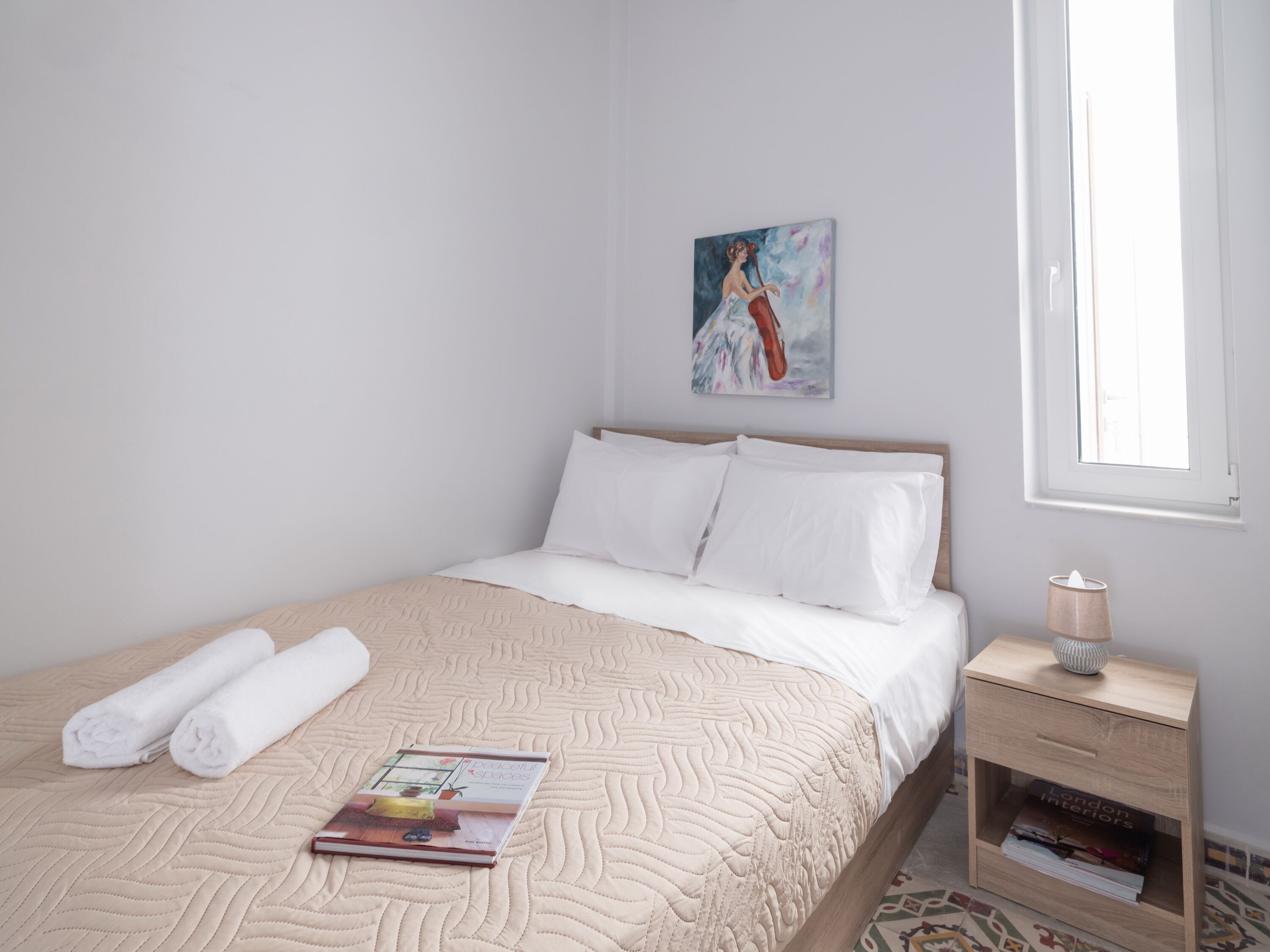 A neatly made bed with white linens and a beige quilt, a book titled "Peaceful Spaces" on the bed, a painting on the wall, and a bedside table with a lamp and a magazine.