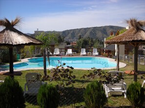 A swimming pool with lounge chairs and thatched umbrellas, surrounded by greenery and hills in the background. - Cabañas Brisa del Lago (Villa Carlos Paz)