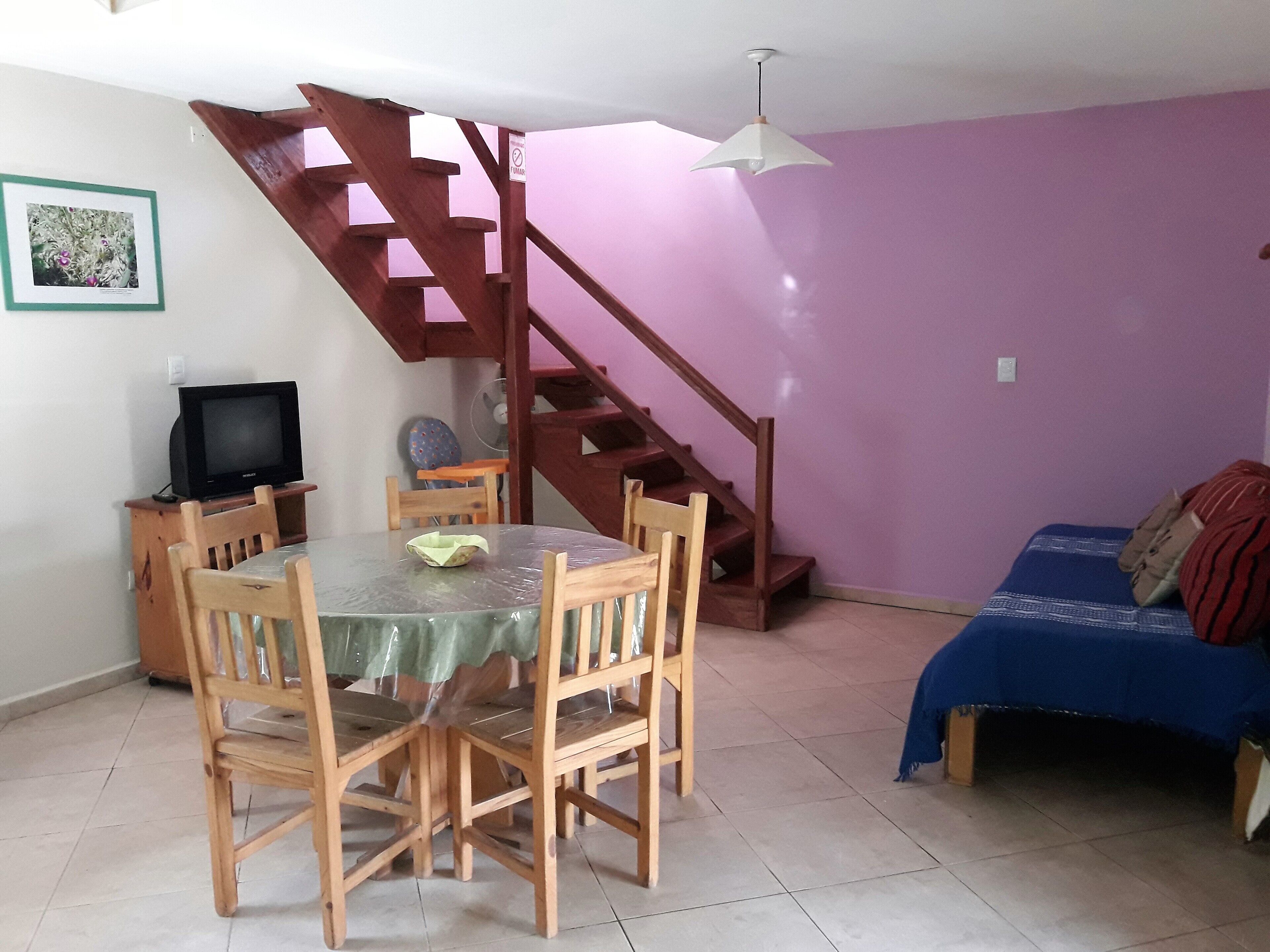 A dining area with a table and chairs, a television on a stand, a staircase, and a bed with a blue cover.