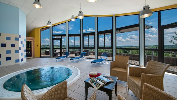 Un vaste espace intérieur abritant une piscine, avec des chaises longues et une petite table, ainsi que de grandes fenêtres offrant une vue sur l’extérieur.