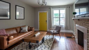 Un salon avec un canapé en cuir marron, un tapis à motifs, une table basse, un fauteuil à motifs, une porte jaune, une cheminée et des tableaux encadrés accrochés au mur.
