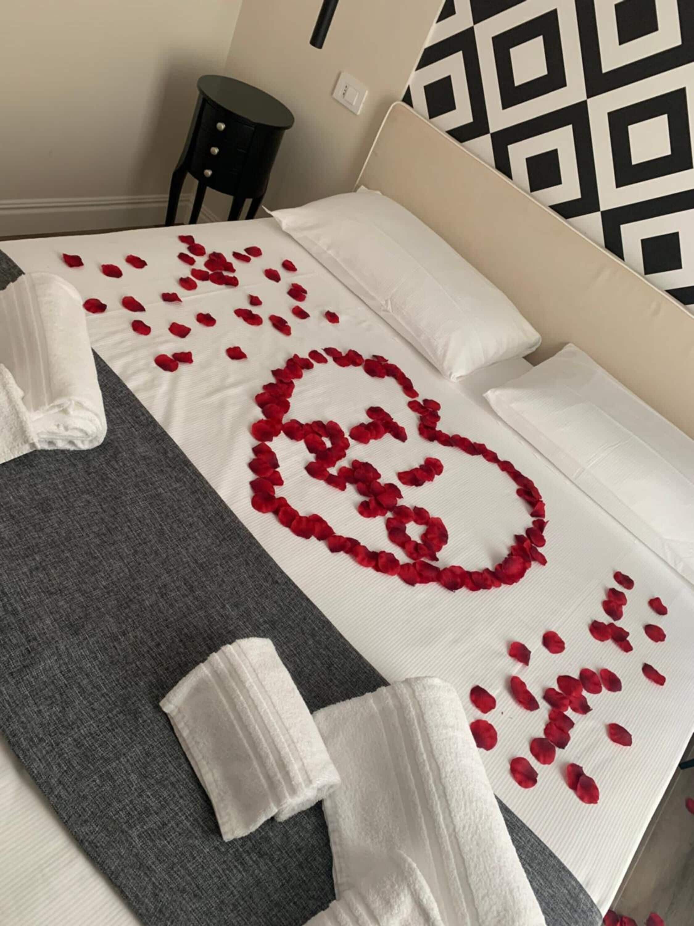 A neatly made bed with rose petals arranged in a heart shape, white towels, and a grey blanket.