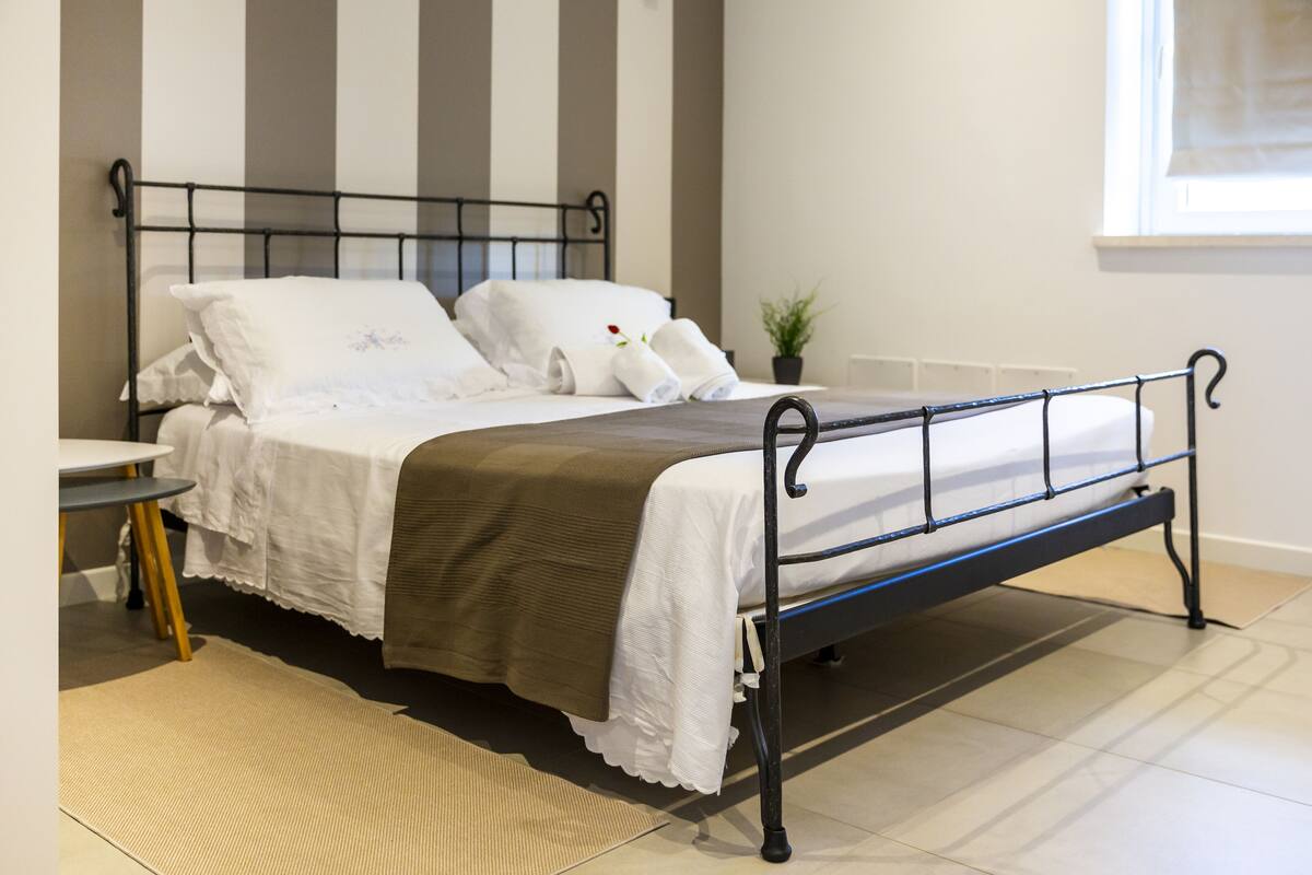 A neatly made bed with white linens and a brown throw, a small table with a yellow base, and a potted plant on the windowsill.