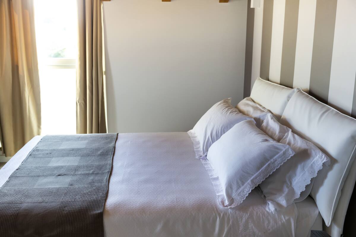 A neatly made bed with white linens and a gray blanket, a striped headboard, and a window with curtains.