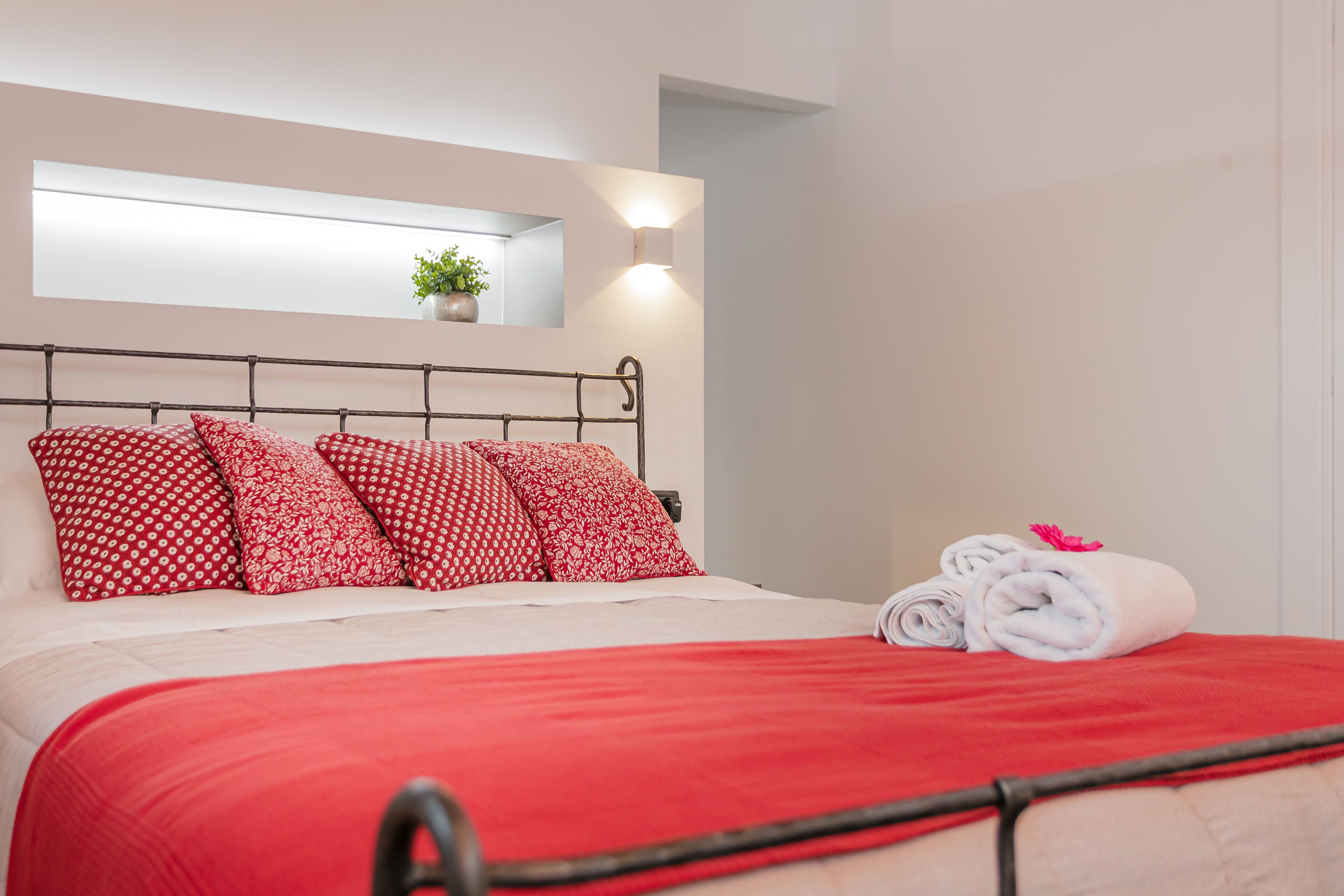 Una cama bien tendida con ropa de cama y almohadas rojas, una pequeña planta en maceta en la pared y una toalla enrollada sobre la cama.