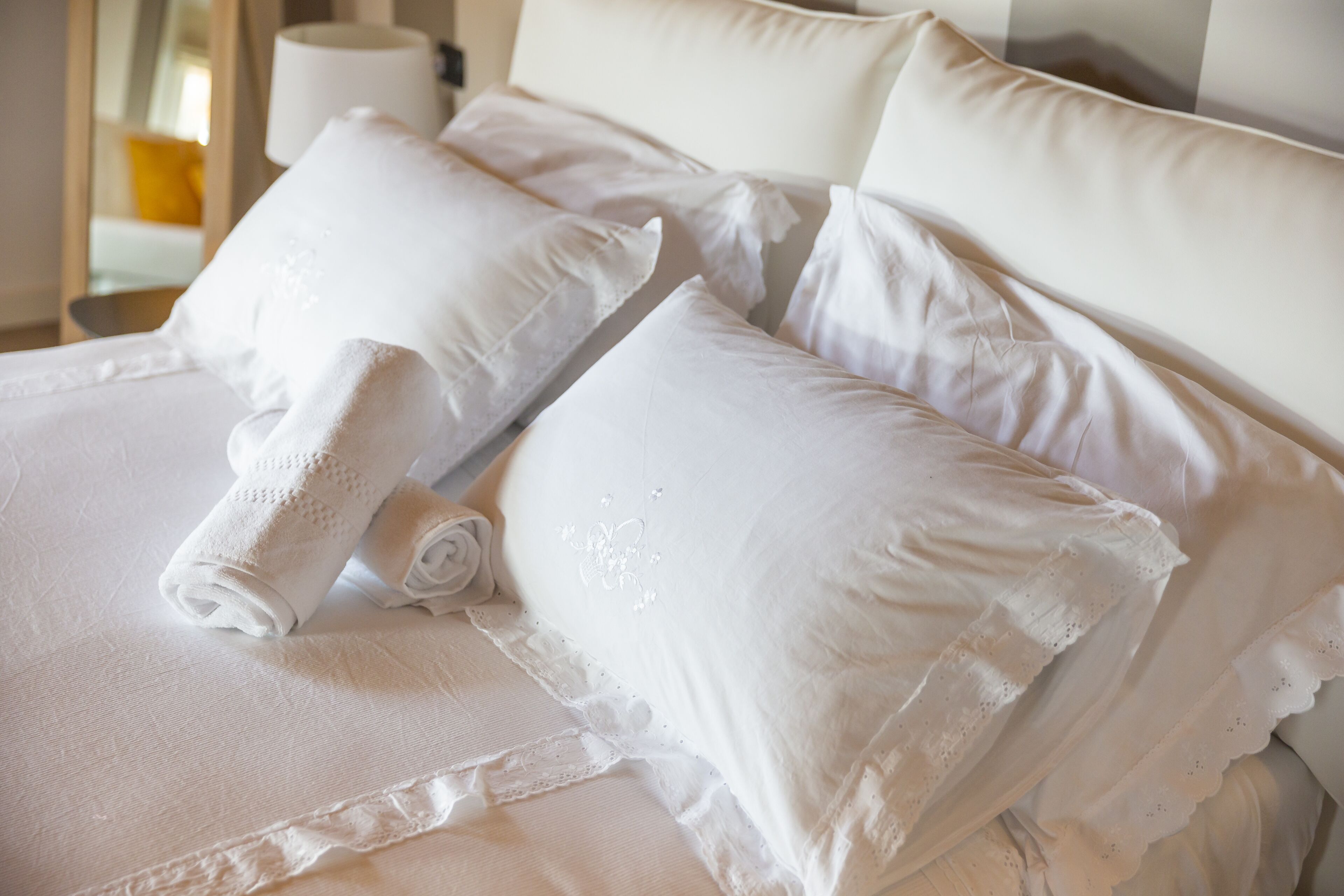 A neatly made bed with white linens and pillows, a rolled towel, and a lamp on a bedside table.