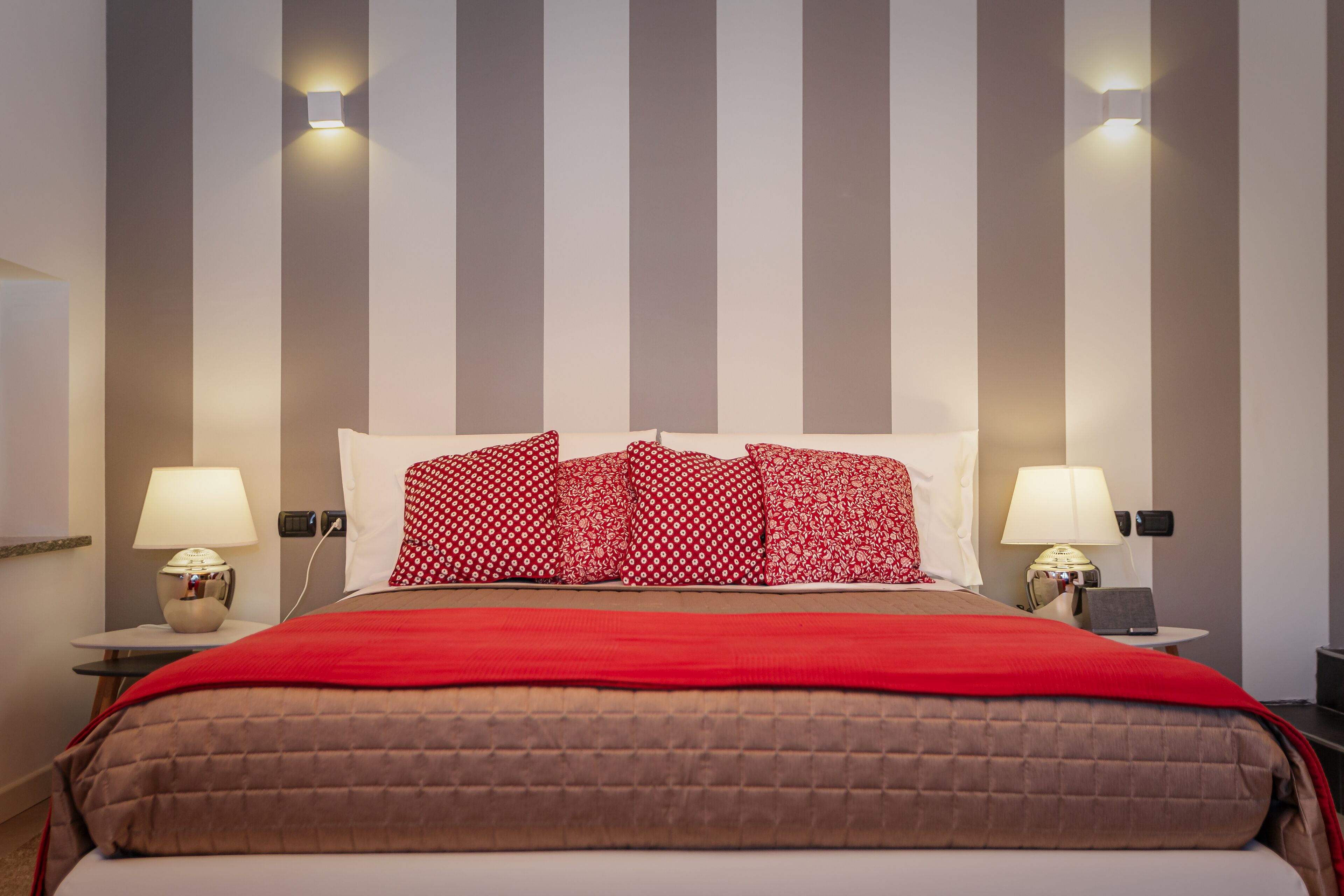 Una habitación de hotel con una cama, almohadas estampadas en rojo y blanco, una manta roja y dos lámparas de noche.