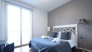Ein Hotelzimmer mit Bett, Nachttisch, Lampe und Blick nach draußen.