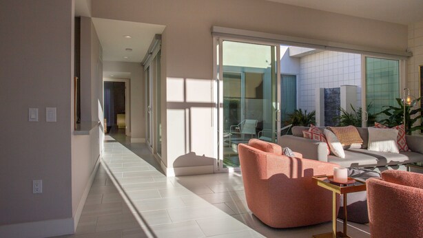 Una sala de estar moderna con un sofá y sillones, un ventanal de vidrio que da a un balcón y un pasillo con piso en tonos claros.