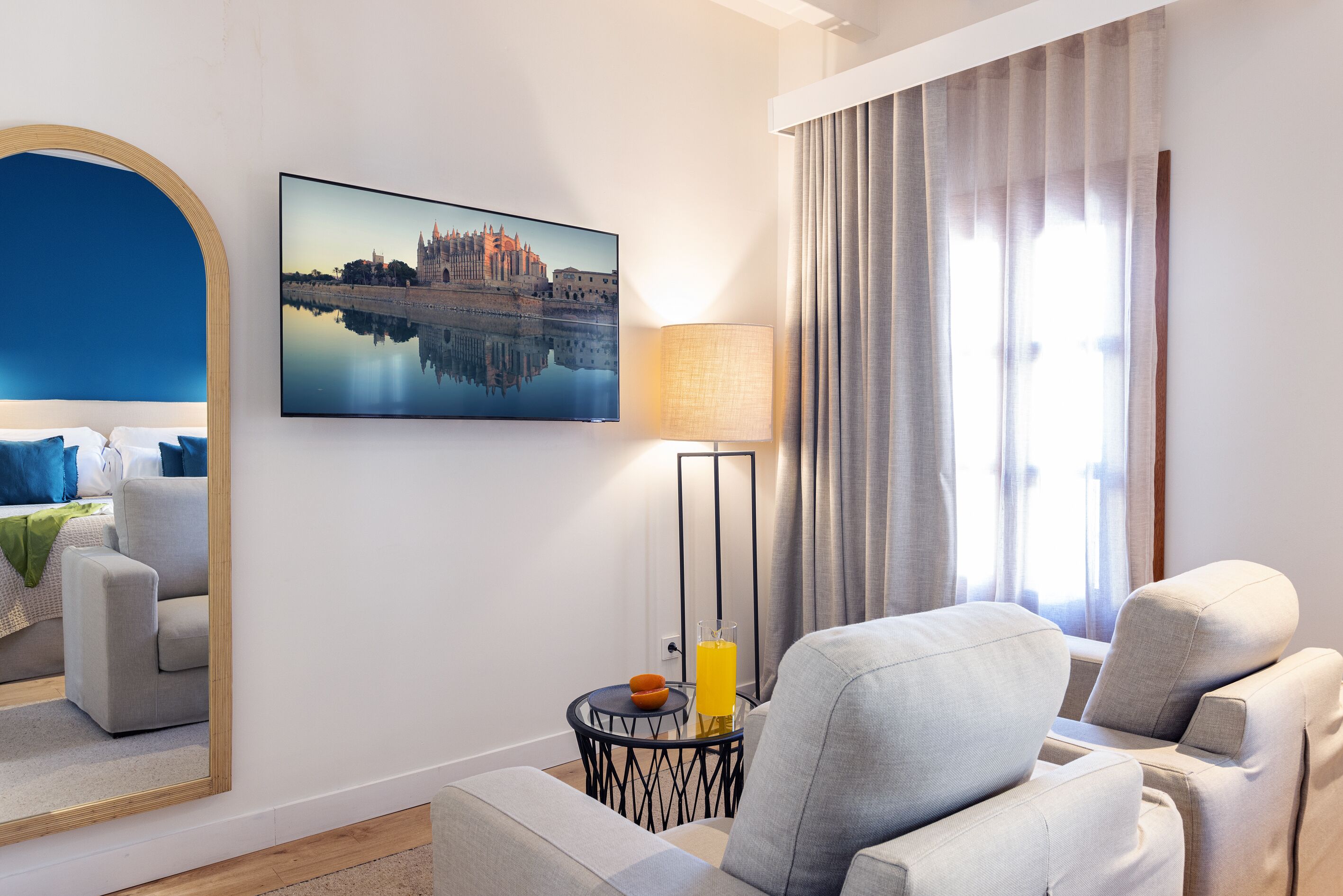 Une chambre d’hôtel moderne avec un grand miroir, un canapé confortable, une petite table avec une lampe et un tableau représentant un paysage urbain accroché au mur.
