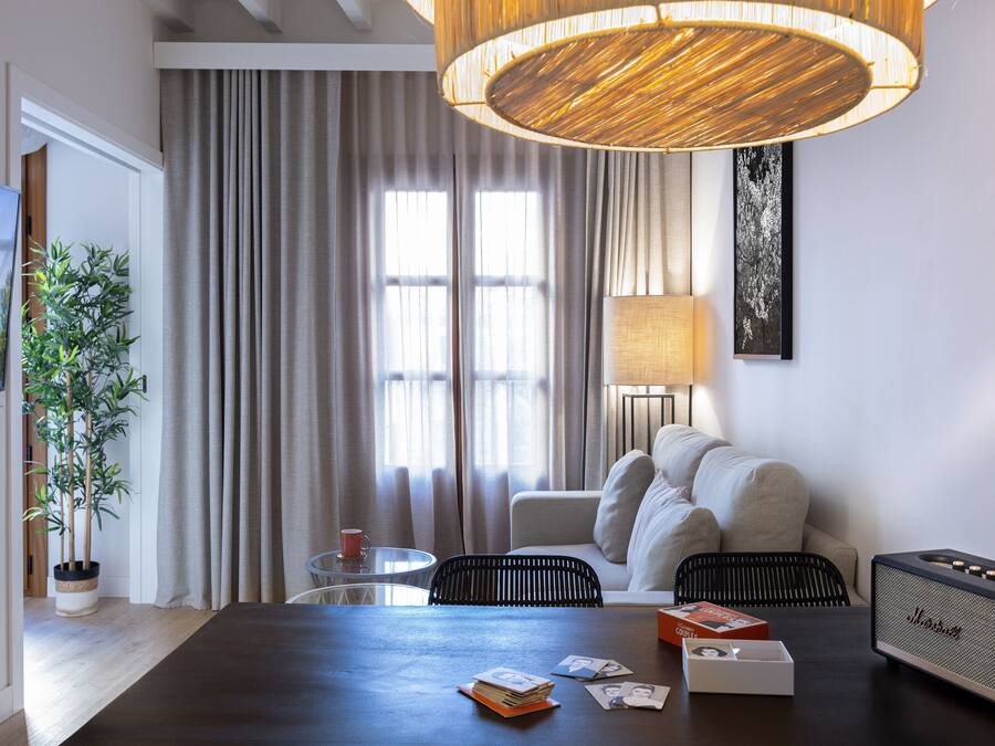 Una habitación de hotel moderna con una mesa de madera oscura, un sofá, una lámpara, un altavoz y un ventanal con cortinas.
