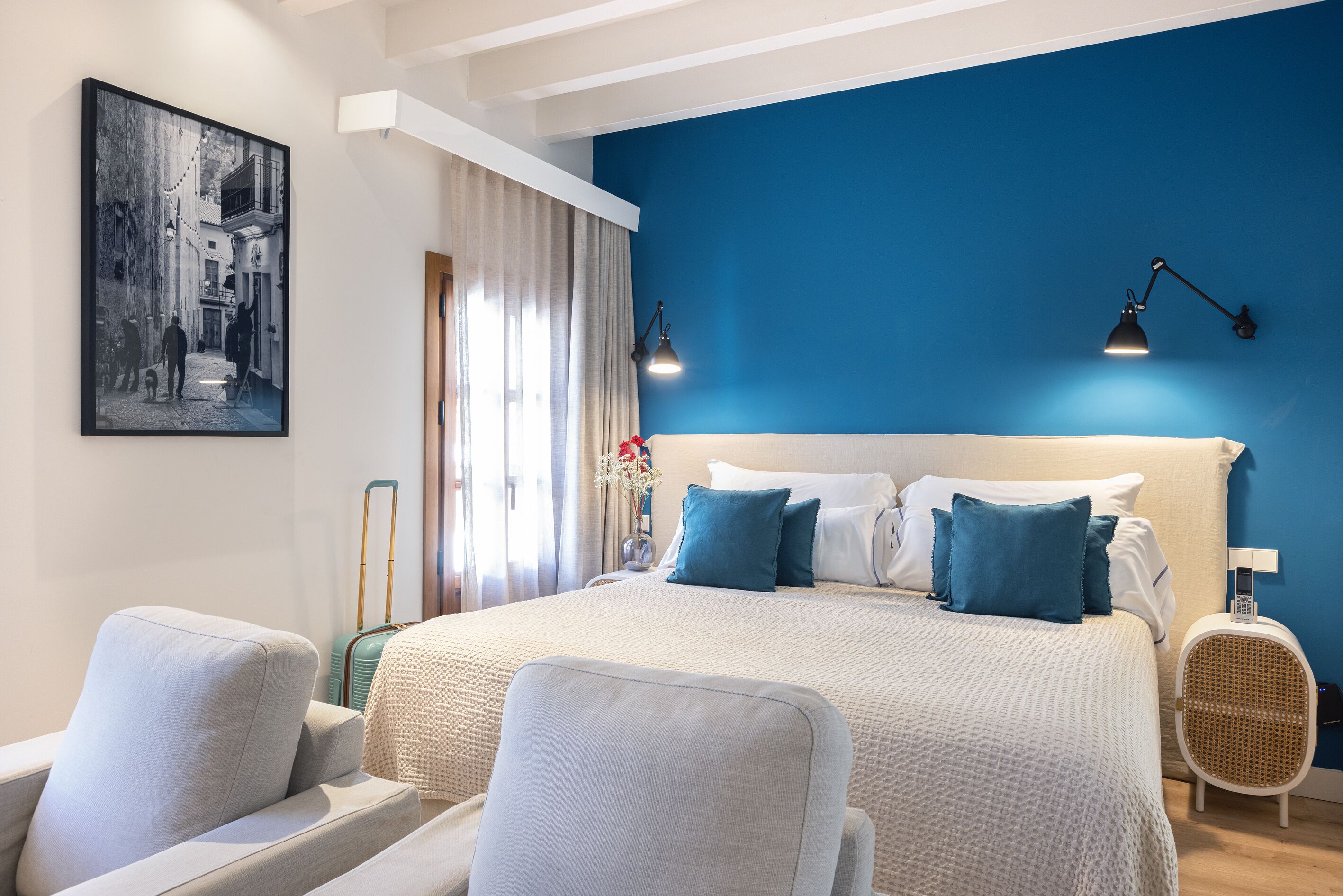 A hotel room with a large bed, two bedside tables, a chair, a wall-mounted lamp, and a framed picture on the wall.
