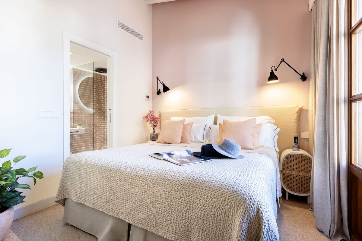 Une chambre à coucher bien aménagée, avec un grand lit, des lampes de chevet, un fauteuil en osier et une petite table sur laquelle est posé un vase.