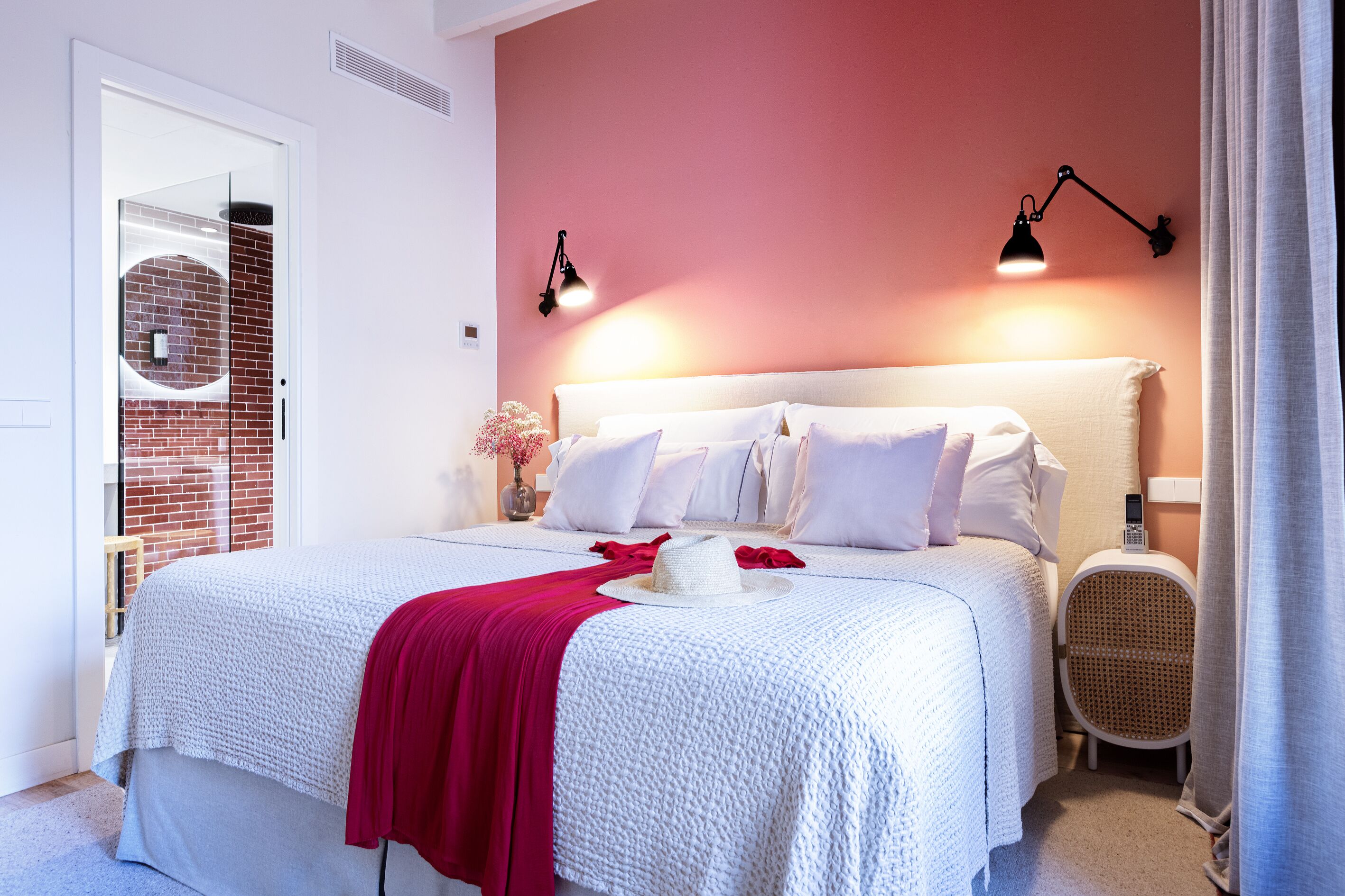 Una cama bien hecha con sábanas blancas y una manta roja, una mesita de noche con una lámpara, y una pared de ladrillo al fondo.