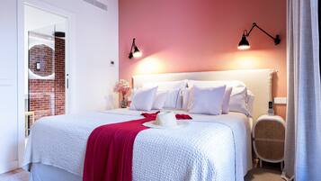 Una cama bien hecha con sábanas blancas y una manta roja, una mesita de noche con una lámpara, y una pared de ladrillo al fondo.