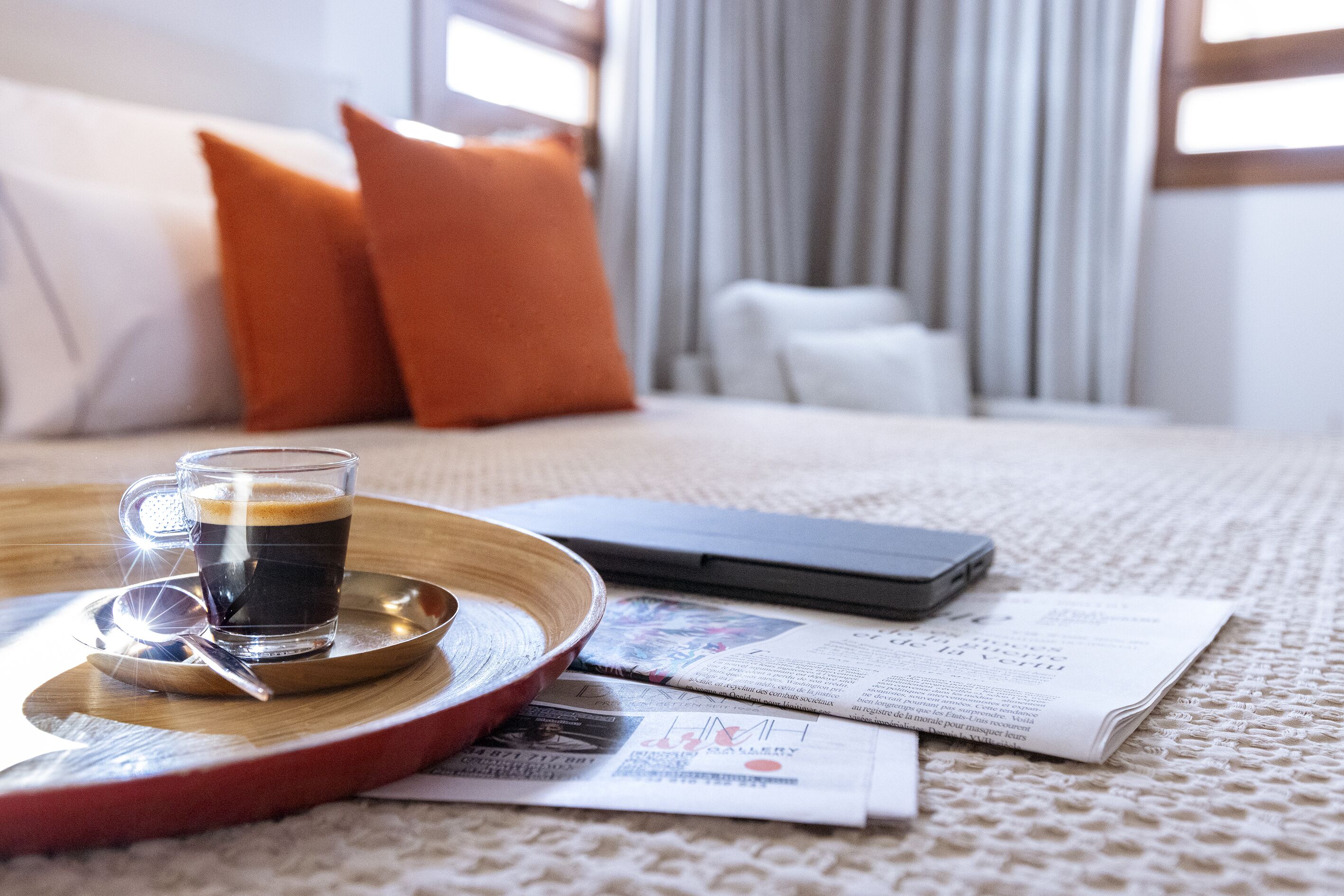 Habitación de hotel ordenada con cama, mesita de noche, taza de café, teléfono inteligente y periódicos.