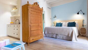 Un dormitorio con un armario de madera, una cama con ropa de cama azul y blanca, una mesita con un libro y una silla de mimbre.