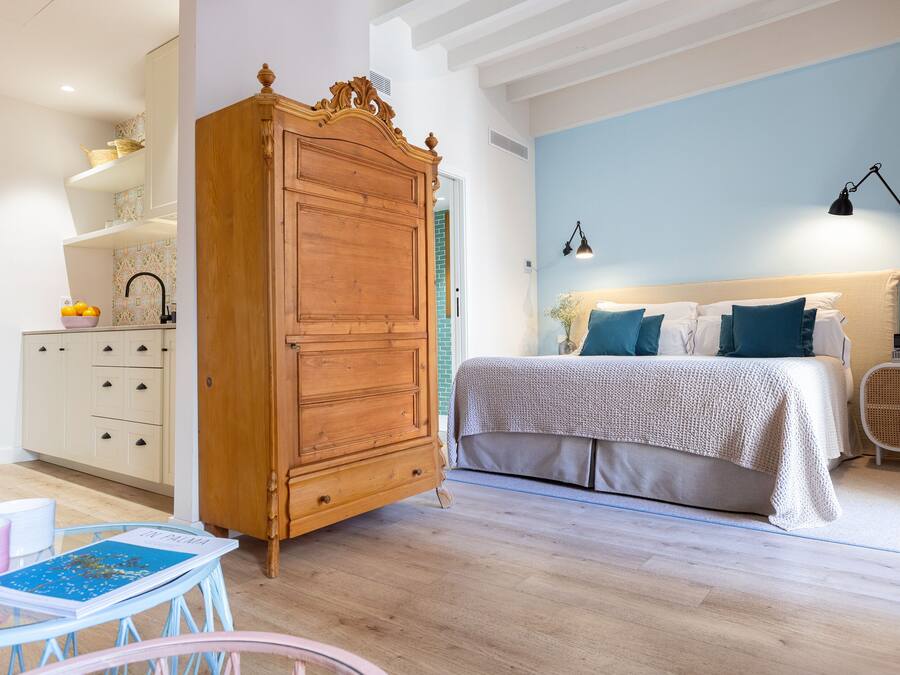 Un dormitorio con un armario de madera, una cama con ropa de cama azul y blanca, una mesita con un libro y una silla de mimbre.