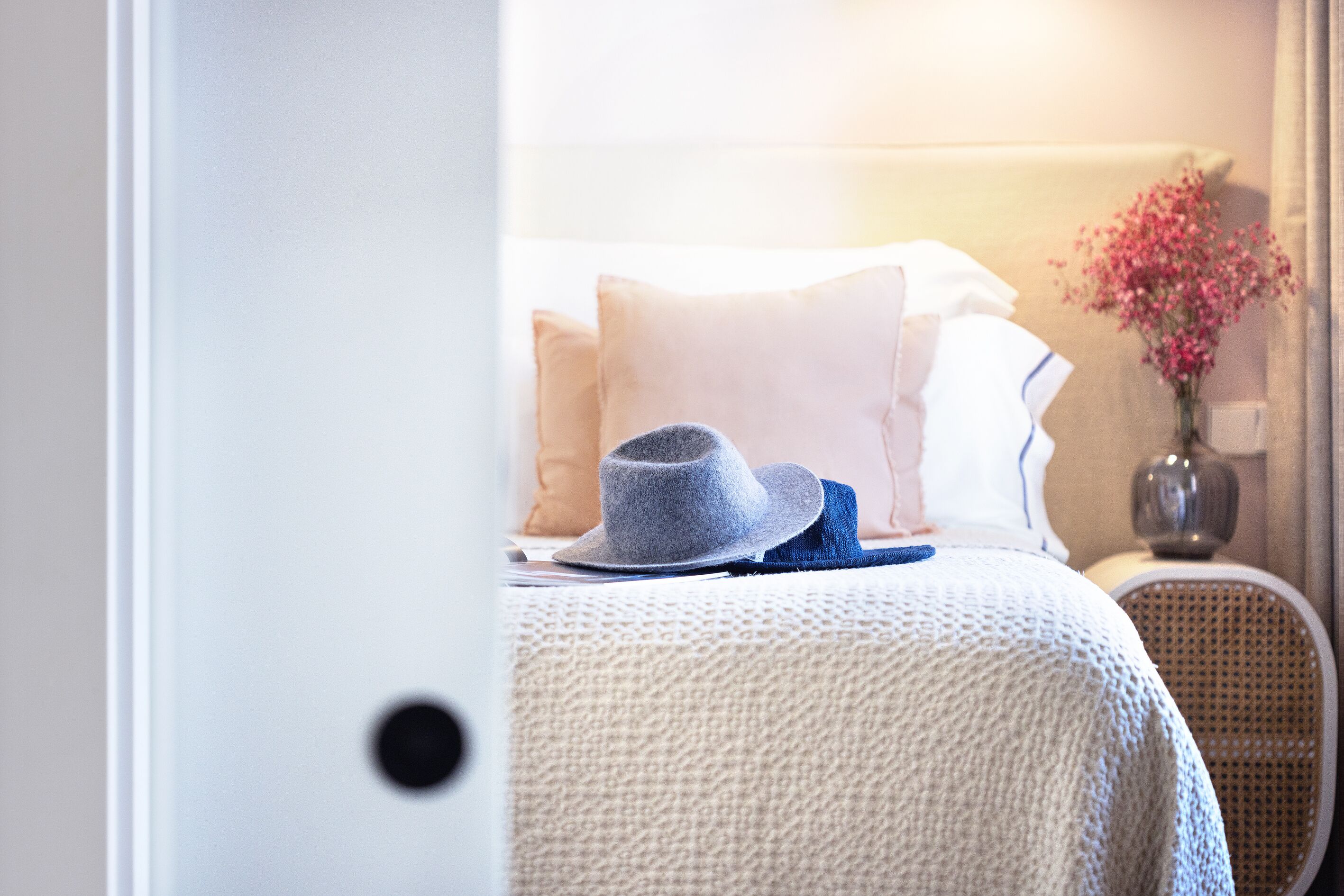 Una cama bien hecha con un sombrero gris y un paño azul encima, una manta blanca de punto y un florero con flores rosas sobre una mesita de noche.