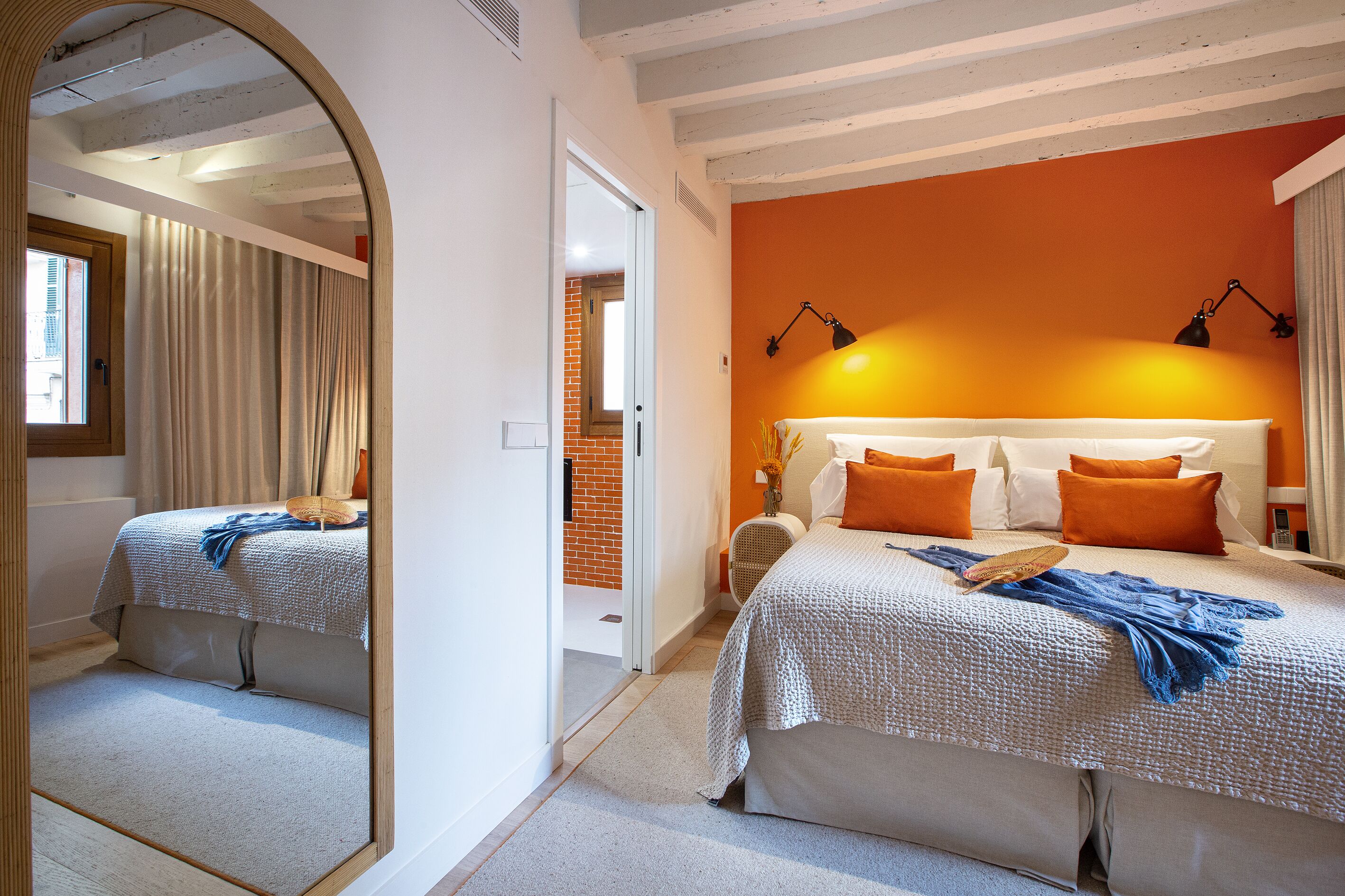 A bedroom with a large mirror, a bed with orange and white bedding, a bedside table with a lamp, and a view of a brick wall and door.