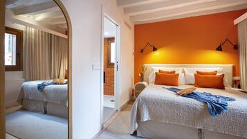 Un dormitorio con un espejo grande, una cama con ropa de cama naranja y blanca, una mesita de noche con una lámpara, y vista a un muro de ladrillo y una puerta.