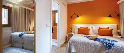 A bedroom with a large mirror, a bed with orange and white bedding, a bedside table with a lamp, and a view of a brick wall and door.