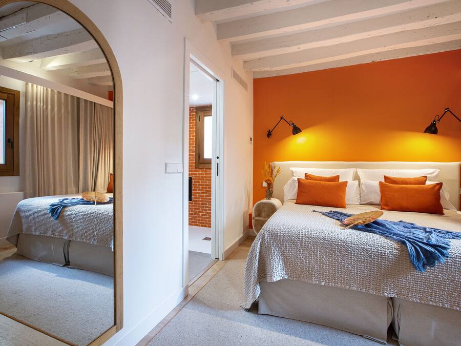 Un dormitorio con un espejo grande, una cama con ropa de cama naranja y blanca, una mesita de noche con una lámpara, y vista a un muro de ladrillo y una puerta.