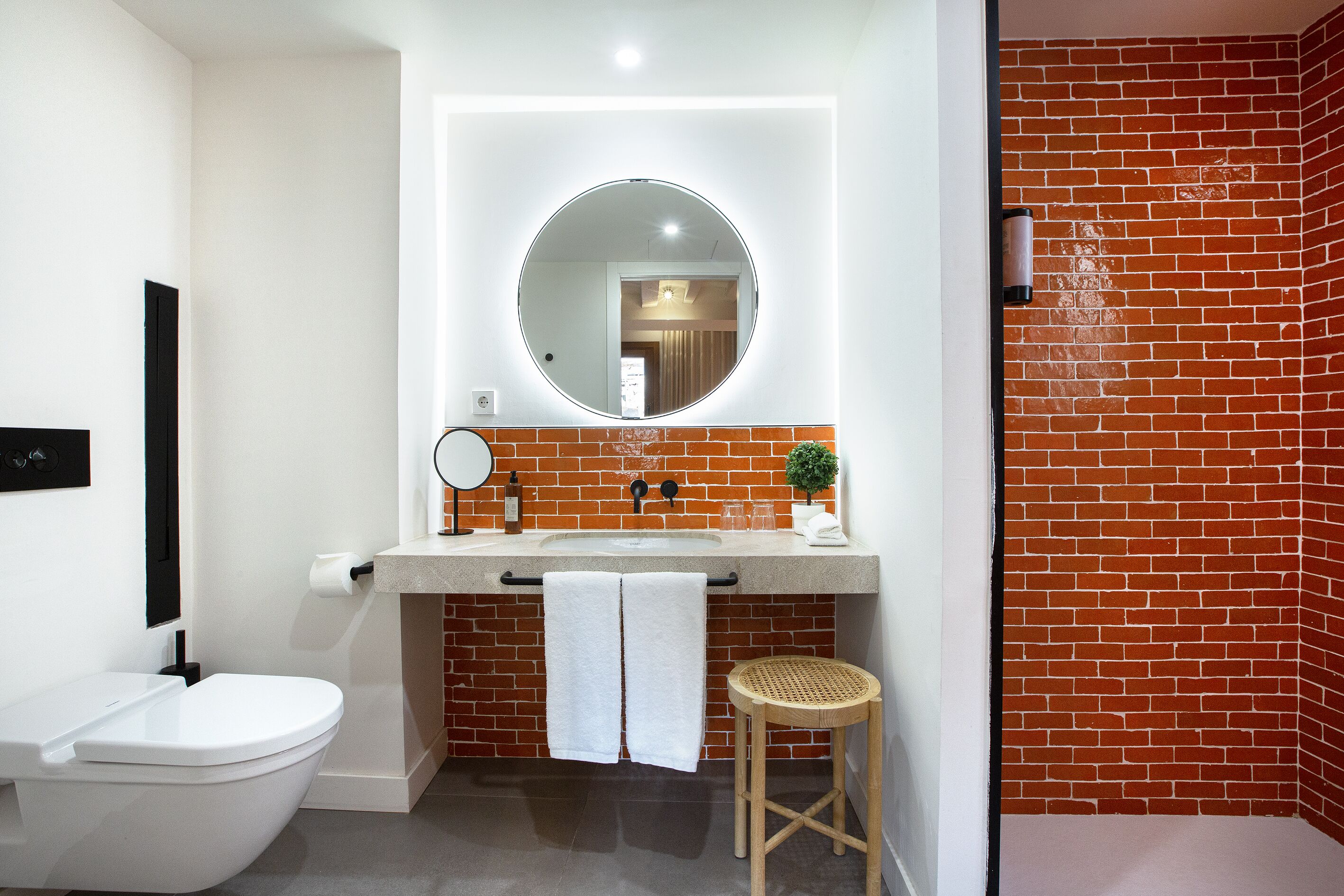 Une salle de bain moderne avec des toilettes blanches, un miroir rond, un lavabo avec un porte-serviettes et un mur en briques.