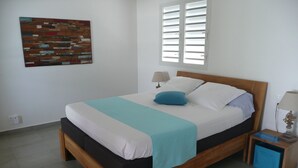 Een net opgemaakt bed met een houten frame, een blauw kussen en een wit-blauwe dekbedovertrek. Er staat een klein nachtkastje met een lamp en een blauwe opbergbox.