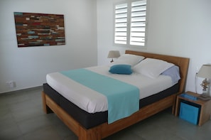 A neatly made bed with a wooden frame, a blue pillow, and a white and blue bedspread. There is a small bedside table with a lamp and a blue storage box. - BUNGALOW HOME PRM. (Poirier - Pigeon Bouillante)