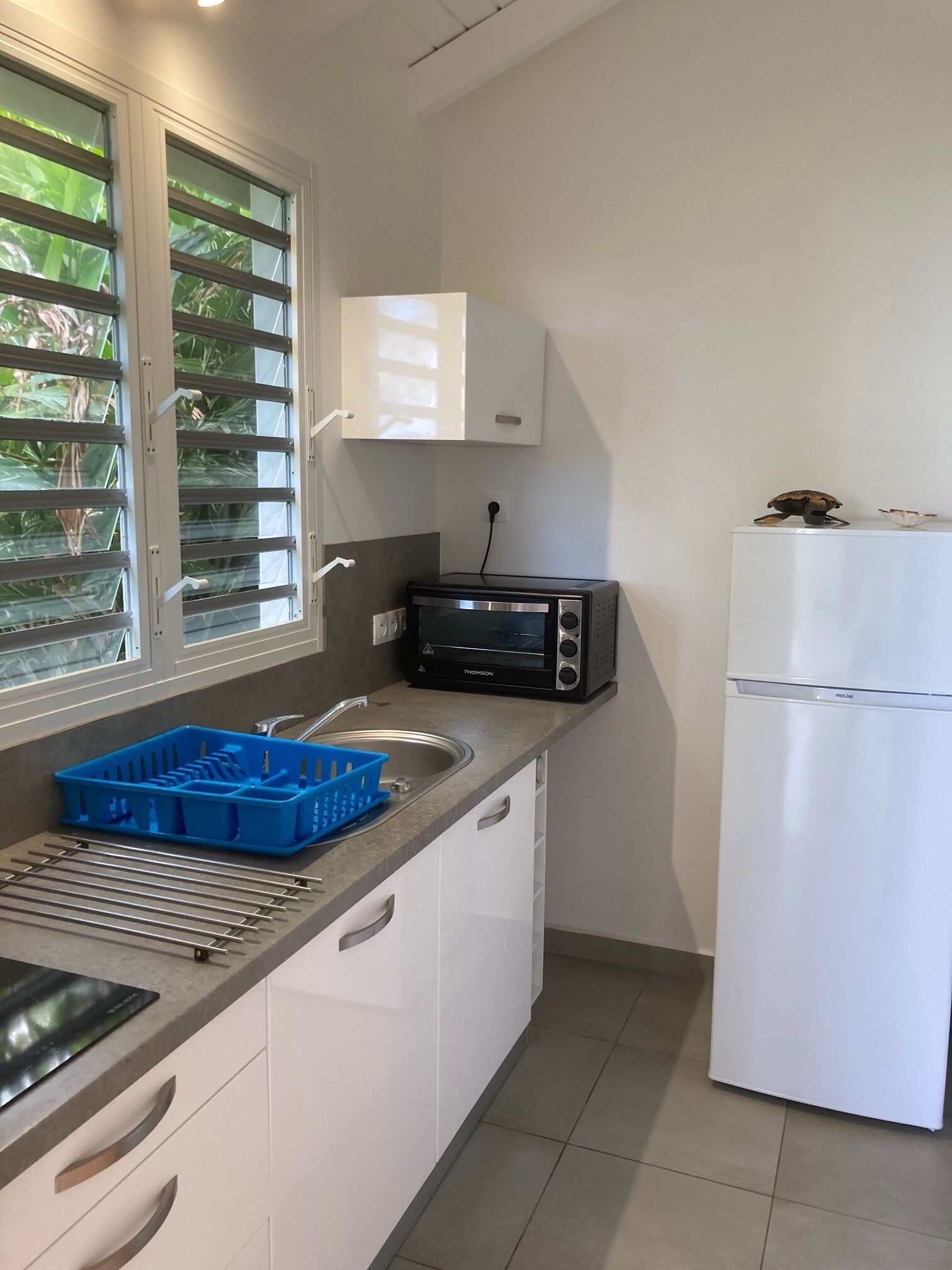 Eine kompakte Küche mit integriertem Mikrowellenherd, Toaster und Kühlschrank. Es gibt ein Fenster mit Jalousien, eine Spüle mit blauem Geschirrgestell und eine Arbeitsfläche mit Geschirrtrockner.
