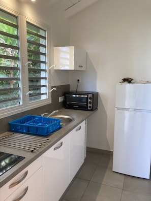 A compact kitchen with a built-in microwave, a toaster, and a refrigerator. There is a window with blinds, a sink with a blue dish rack, and a countertop with a dish drying rack. - BUNGALOW HOME PRM. (Poirier - Pigeon Bouillante)