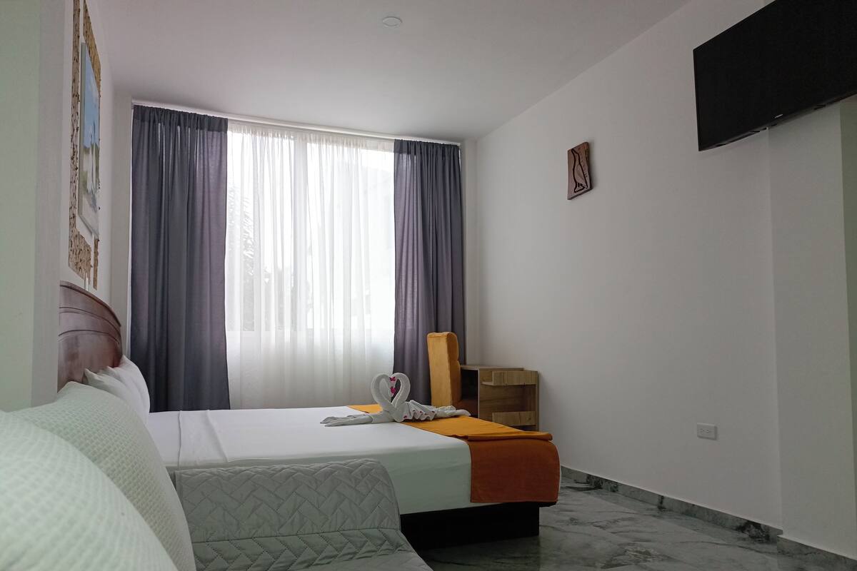 Ein Hotelzimmer mit zwei Betten, einem Holfnachtkästchen, einem Flachbildfernseher und einem großen Fenster mit Vorhängen.