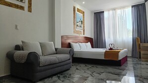 Habitación de hotel con cama, sofá, cabecera de madera, piso de mármol y ventana con cortinas.