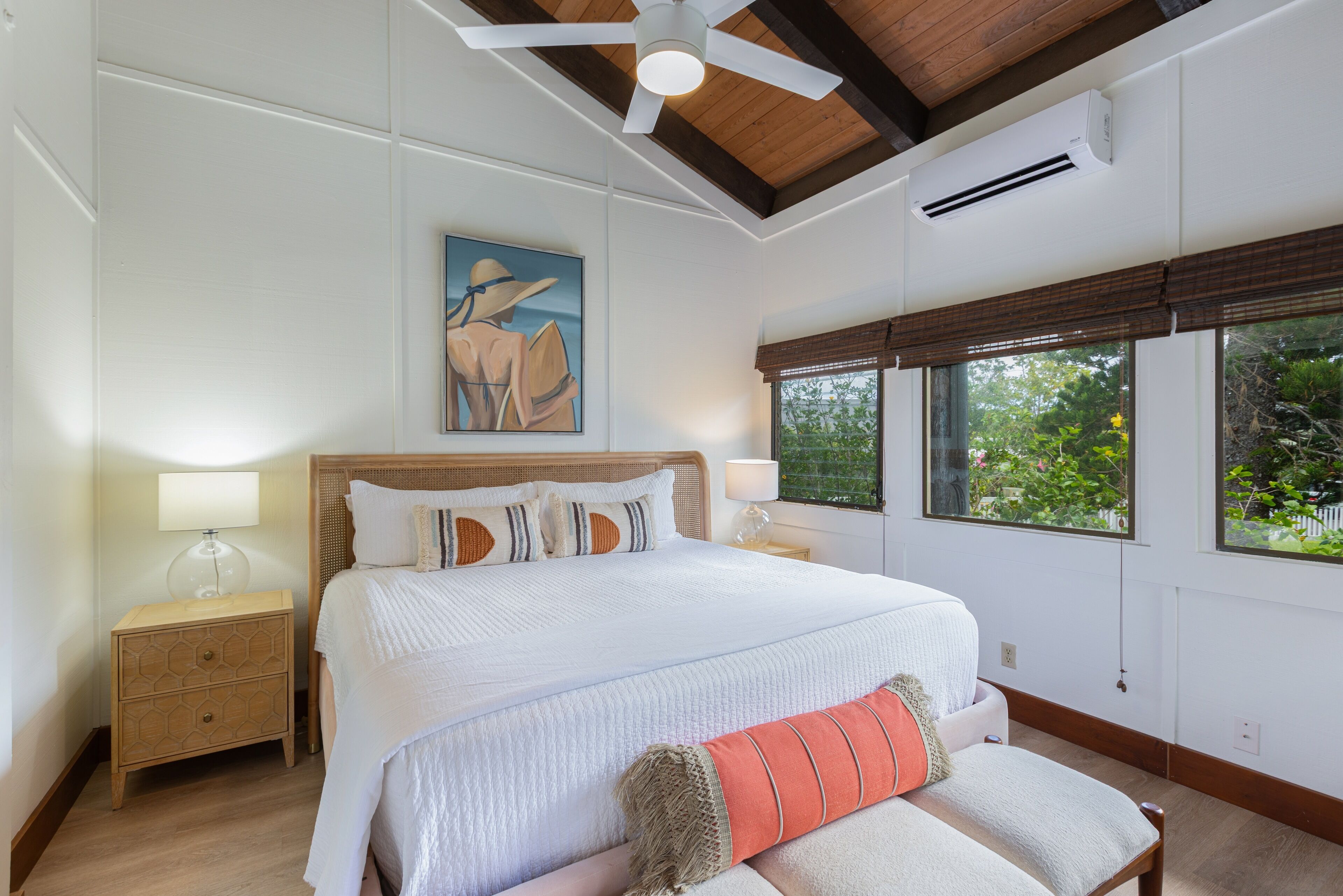 A bedroom with a large bed, wooden headboard, two bedside tables with lamps, a ceiling fan, and a window with a view of greenery.
