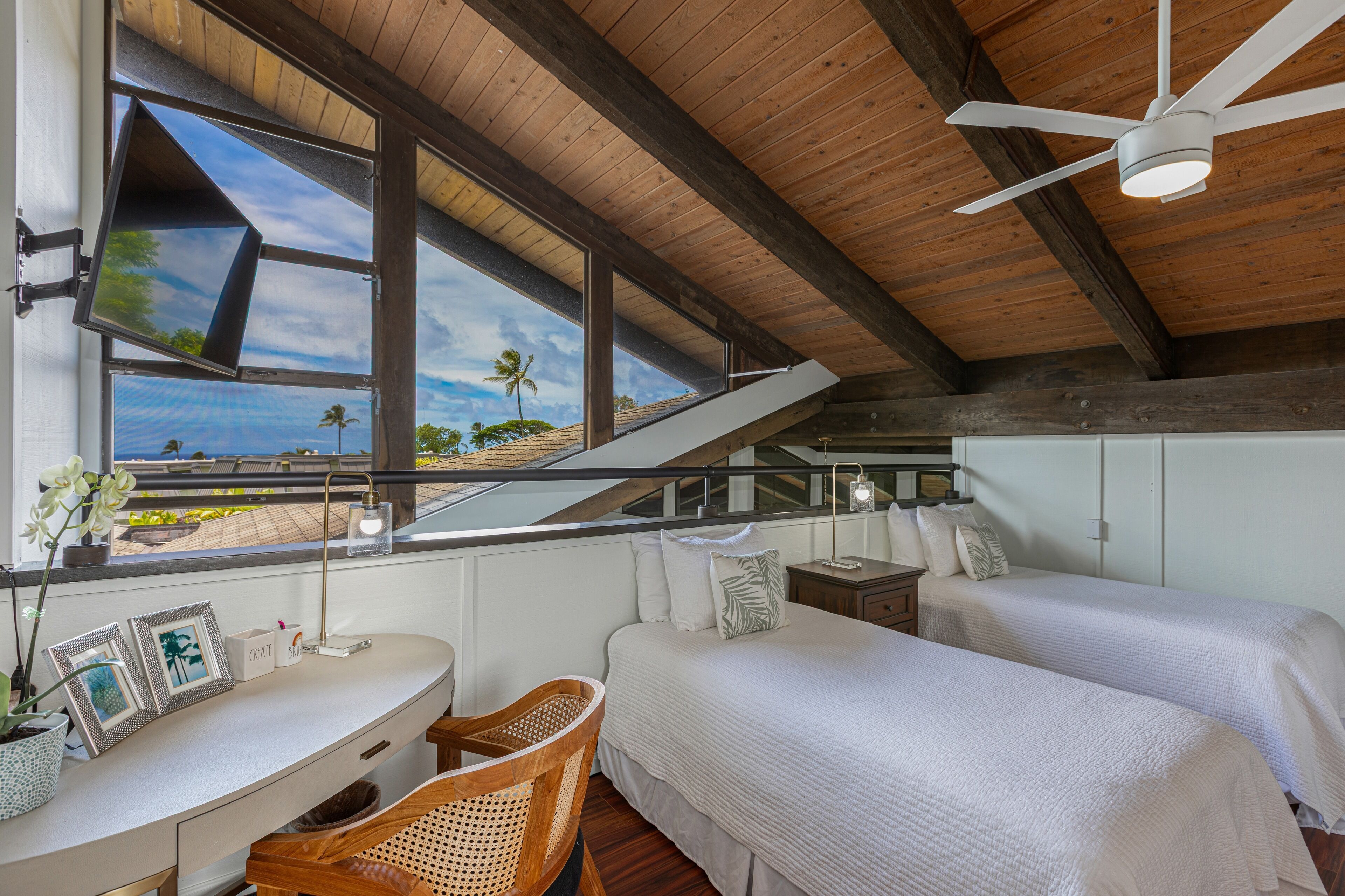 Un dormitorio con dos camas, un escritorio y una silla. Se ve una ventana con vistas a palmeras y al cielo.