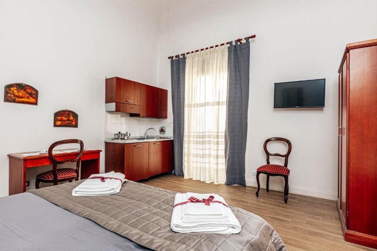 Quarto compacto com cama, escrivaninha, cadeira, pequeno espaço de cozinha, televisão e guarda-roupa.