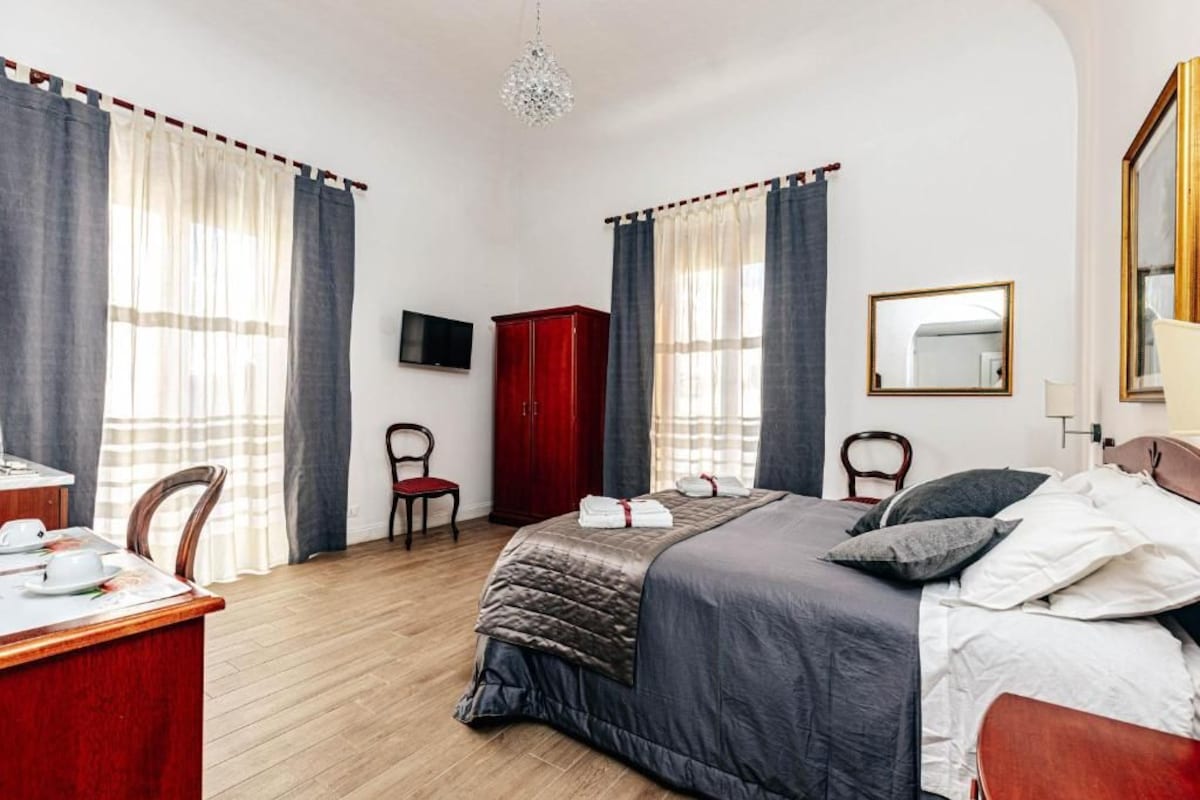 A hotel room with a bed, a desk with a chair, a wardrobe, a television, and a mirror.