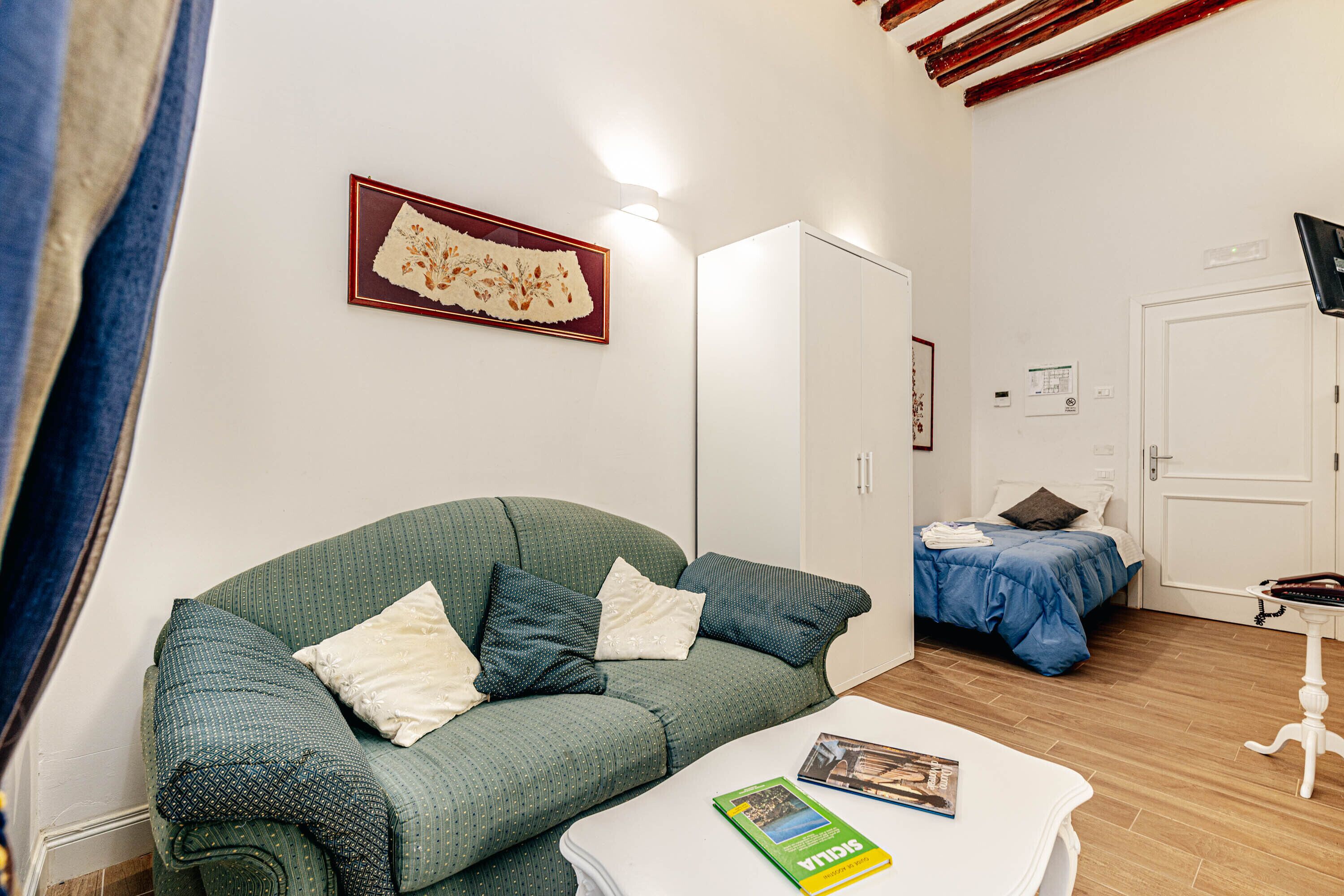 A room with a green sofa, a white table with a book, a bed with a blue blanket, and a white wardrobe.
