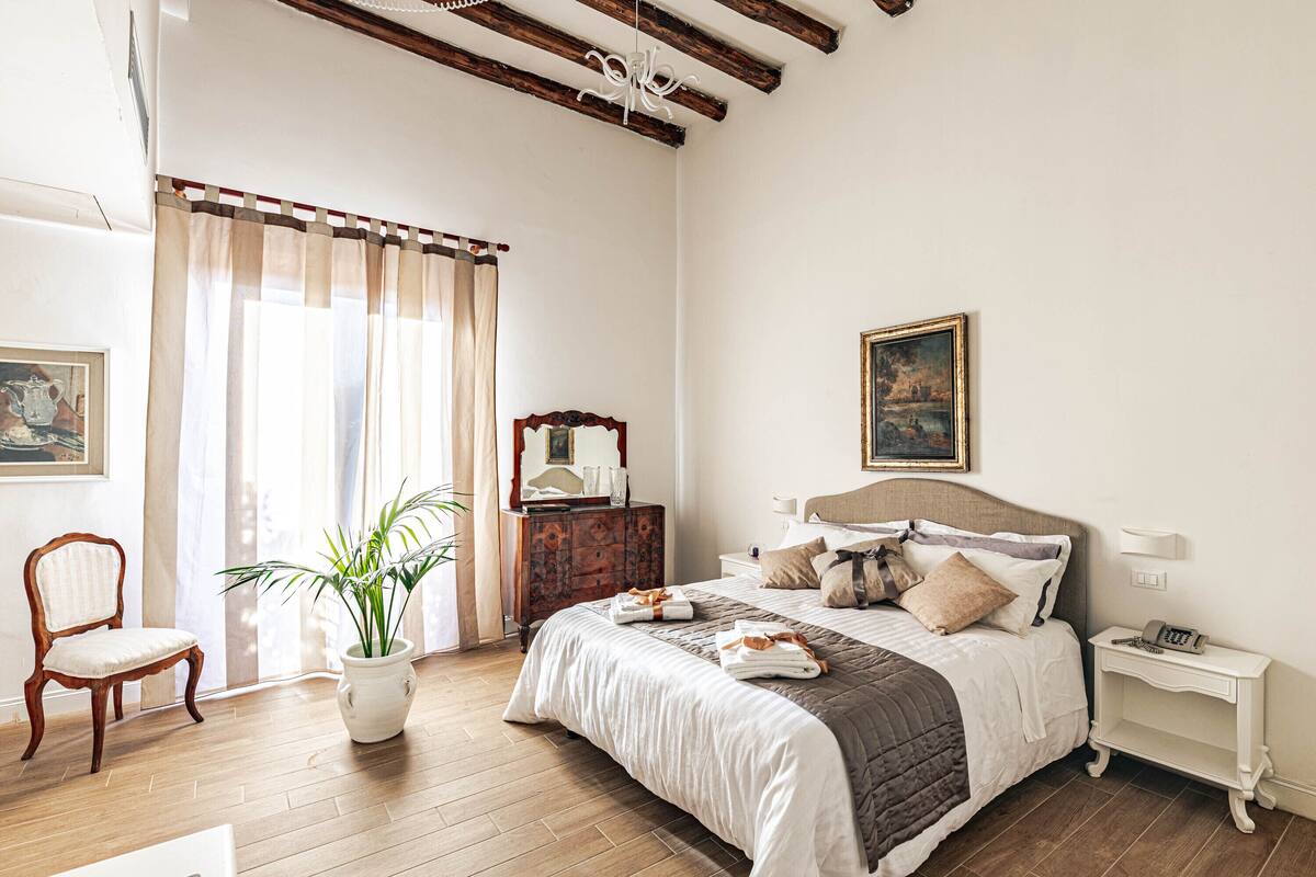 A bedroom with a wooden floor, a bed with a white and brown bedspread, a bedside table with a lamp, a wooden chair, and a potted plant.