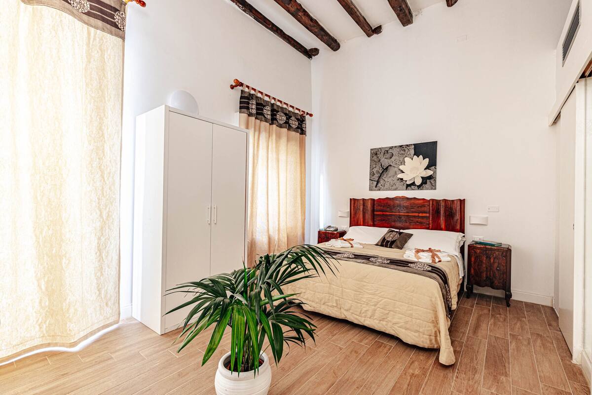 A bedroom with a bed, a nightstand, a wooden headboard, a potted plant, a wardrobe, and a window with curtains.