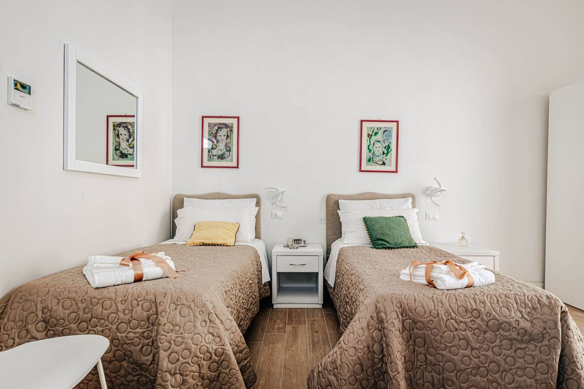 A hotel room with two beds, each with a brown patterned bedspread, white pillows, and a small bedside table with a lamp. There are framed pictures on the wall above each bed.