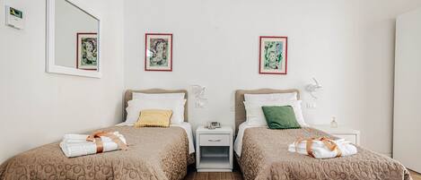 A hotel room with two beds, each with a brown patterned bedspread, white pillows, and a small bedside table with a lamp. There are framed pictures on the wall above each bed.