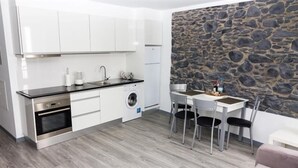 A compact kitchen with white cabinets, a built-in oven, a washing machine, and a stone wall. There is a dining table with chairs and a small white table. - Apartamentos Funchal by Petit Hotels (Funchal)