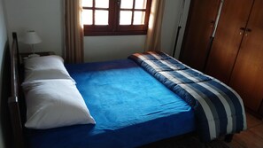 Un lit simple avec une couvre-lit bleue et des oreillers blancs, une armoire en bois et une fenêtre avec des rideaux.