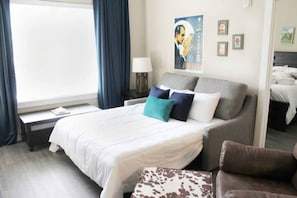Quarto de hotel com uma cama grande, mesa de cabeceira, abajur, um cartaz na parede e uma janela com cortinas.
